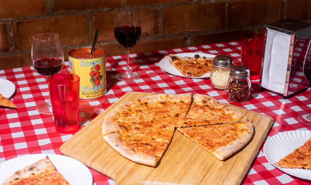 A New York style pizza topped with cheese and tomato on a wooden pizza peel with two hands taking a slice each. Made using a New York-style Pizza Dough recipe.