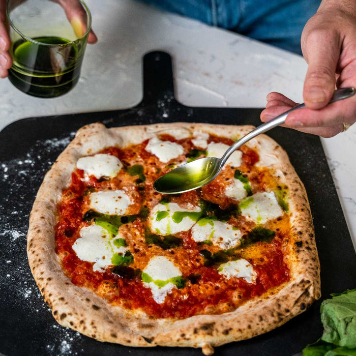 Basil oil being poured out of a glass onto a spoon then drizzled on a pizza topped with tomato sauce and mozzarella