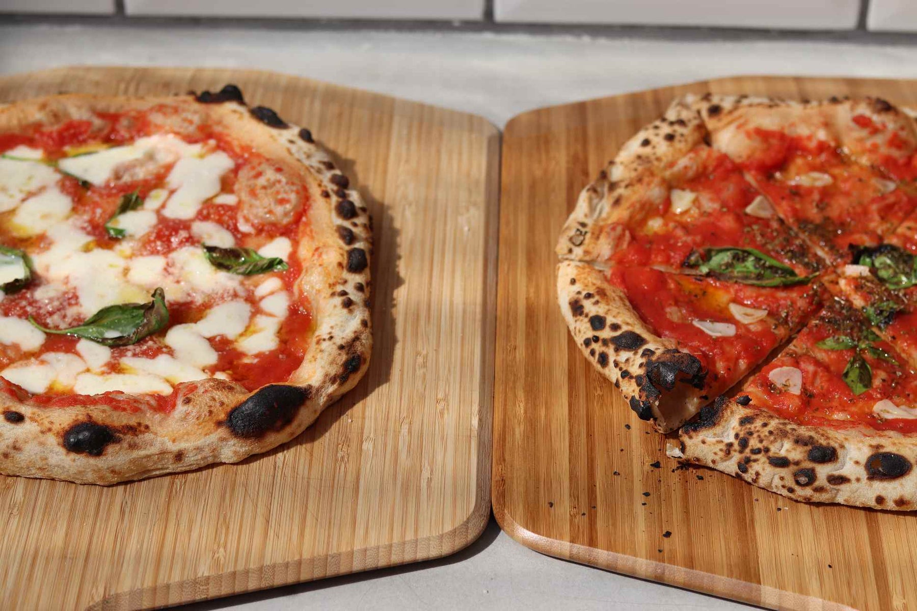 One cooked Margherita biga dough pizza with mozzarella, tomato sauce and basil on an Ooni Bamboo Peel & Serving Board next to a cooked marinara biga dough pizza with tomato sauce, garlic and basil on an Ooni Bamboo Peel & Serving Board.