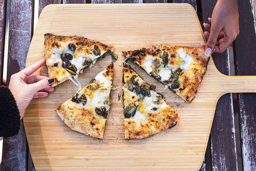 Two hands holding slices of cooked calzone with escarole and provola on top of an Ooni Bamboo Pizza Peel & Serving Board on a table.