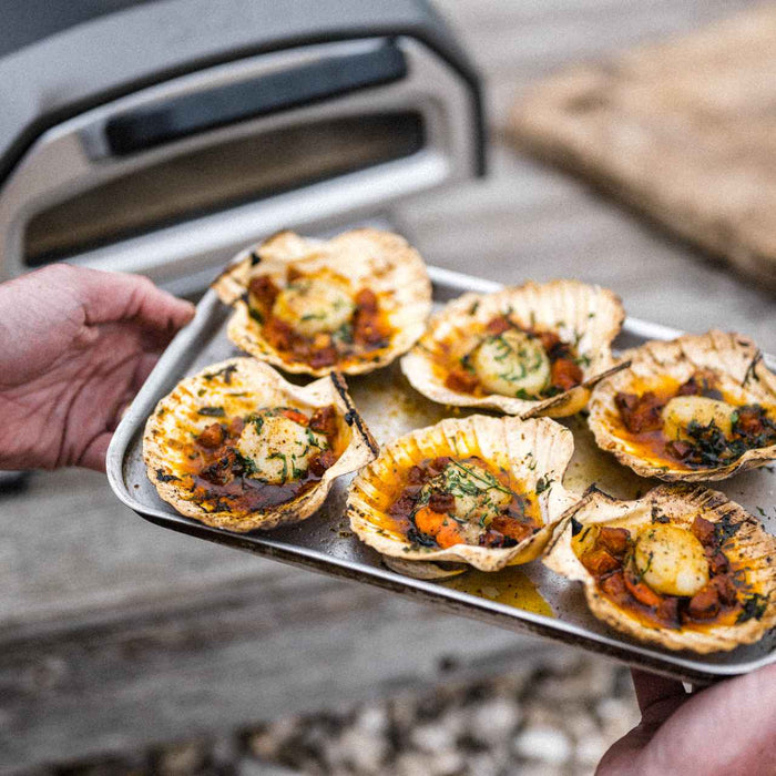 Two hands holding a roasting tray with roasted hand-dived scallops with chorizo, garlic and parsley.