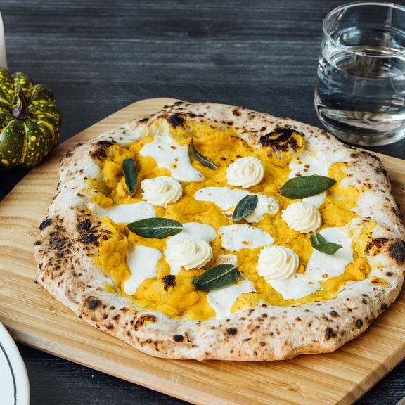 Cooked butternut squash, ricotta and fried sage pizza on an Ooni Bamboo Pizza Peel & Serving Board on a table next to plates, a glass of water and decorative gourds.