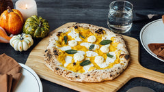 Cooked butternut squash, ricotta and fried sage pizza on an Ooni Bamboo Pizza Peel & Serving Board on a table next to plates, a glass of water and decorative gourds.