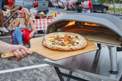 Cooked Philly cheesesteak pizza with cème fraîche, mozzarella, cooked ribeye, provolone, green capsicums and onions on an Ooni Bamboo Pizza Peel & Serving Board next to an Ooni Professional Pizza Cutter Wheel.