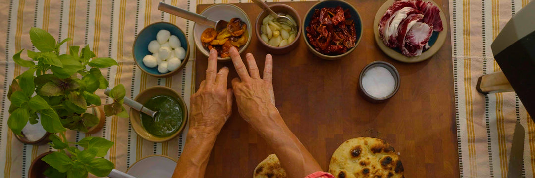 Two hands touching a bowl of marinated baby bell peppers next to bowls of basil pesto, mozzarella balls, garlic confit, capsicums, radicchio and salt.