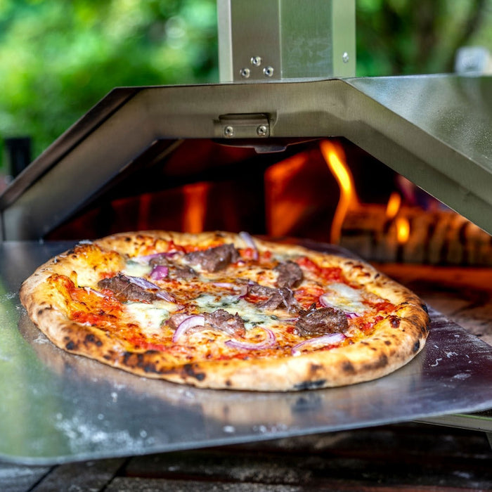 A pizza with topped with steak, blue cheese and red onion on a metal pizza peel being inserted into an Ooni pizza oven. Made using a steak pizza recipe.