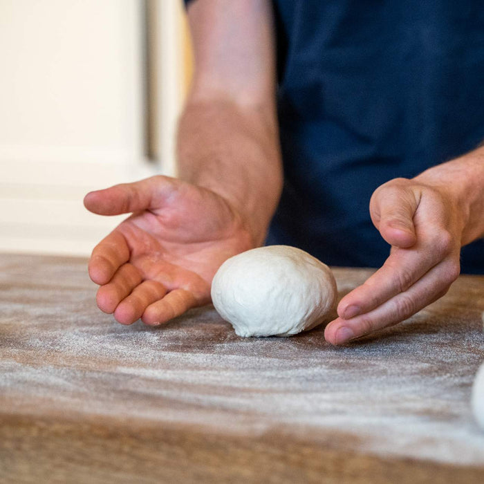 Ball of pizza dough being rolled out on a wooden chopping board, made using a Cold Proof Pizza Dough recipe.