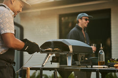 person removing cast iron cookware from Ooni Koda 2. outdoor cooking set up with modular tables, social scene in background. 